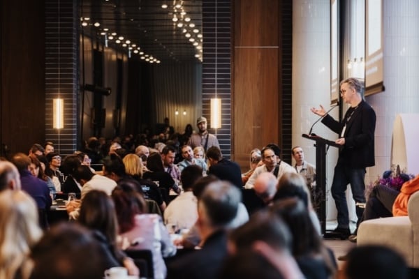 A speaker addresses a full room at a professional event. Attendees sit at round tables, listening closely in a modern, well-lit conference space.
